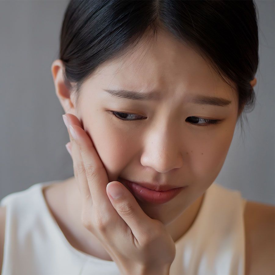 Woman touching her cheek in pain, looking distressed against neutral background.