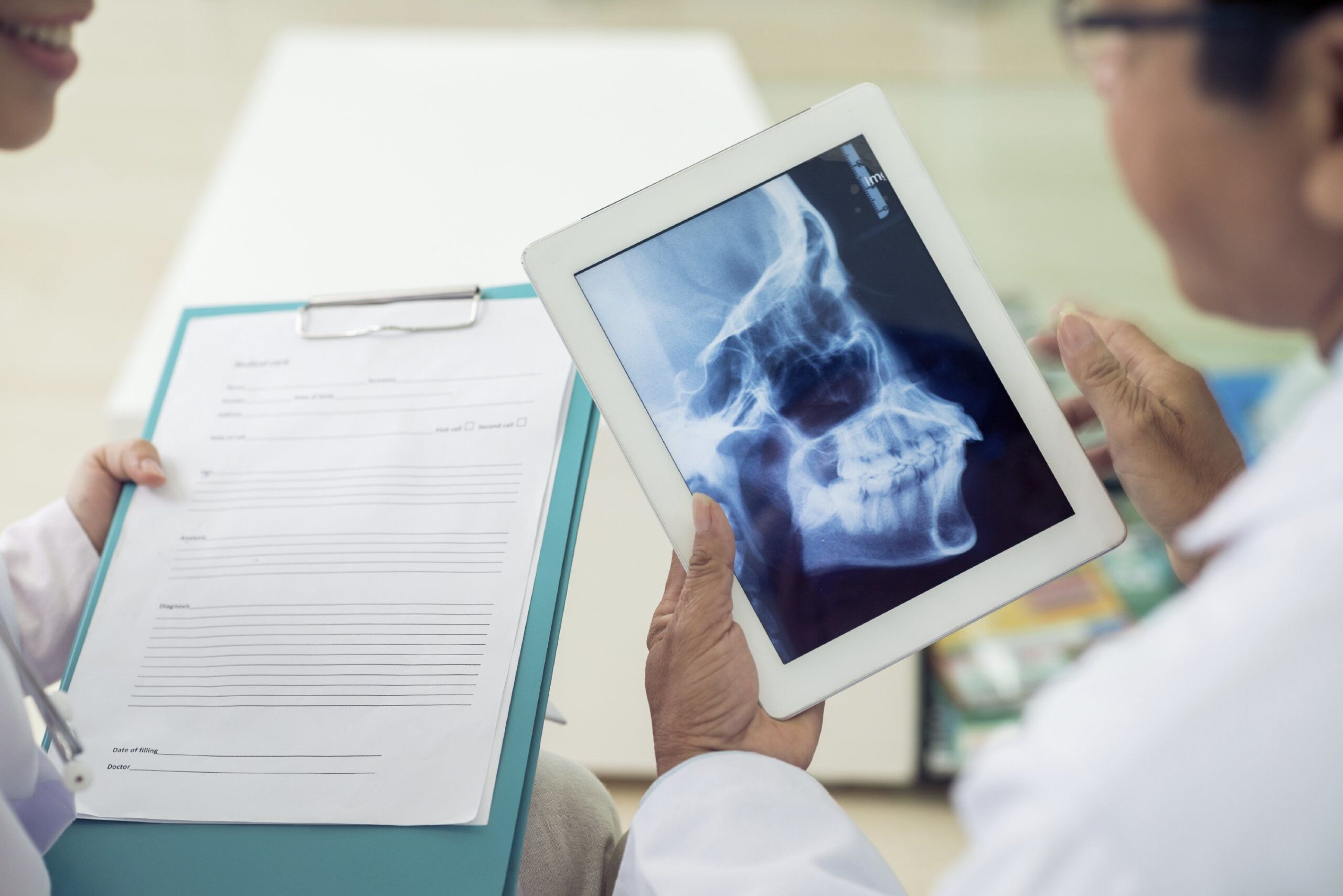 Two dentists examine dental X-ray on tablet while reviewing patient records at desk.