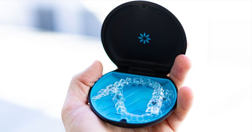 Hand holding open black case revealing blue dental retainer or aligners against blurred background