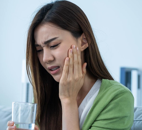 Woman holding cheek in pain, holding water glass, experiencing dental discomfort indoors.