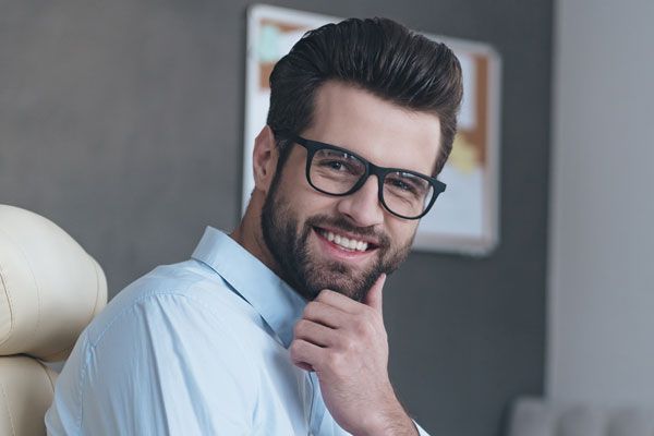 Professional male dentist wearing glasses and blue shirt in modern dental office setting, smiling confidently.
