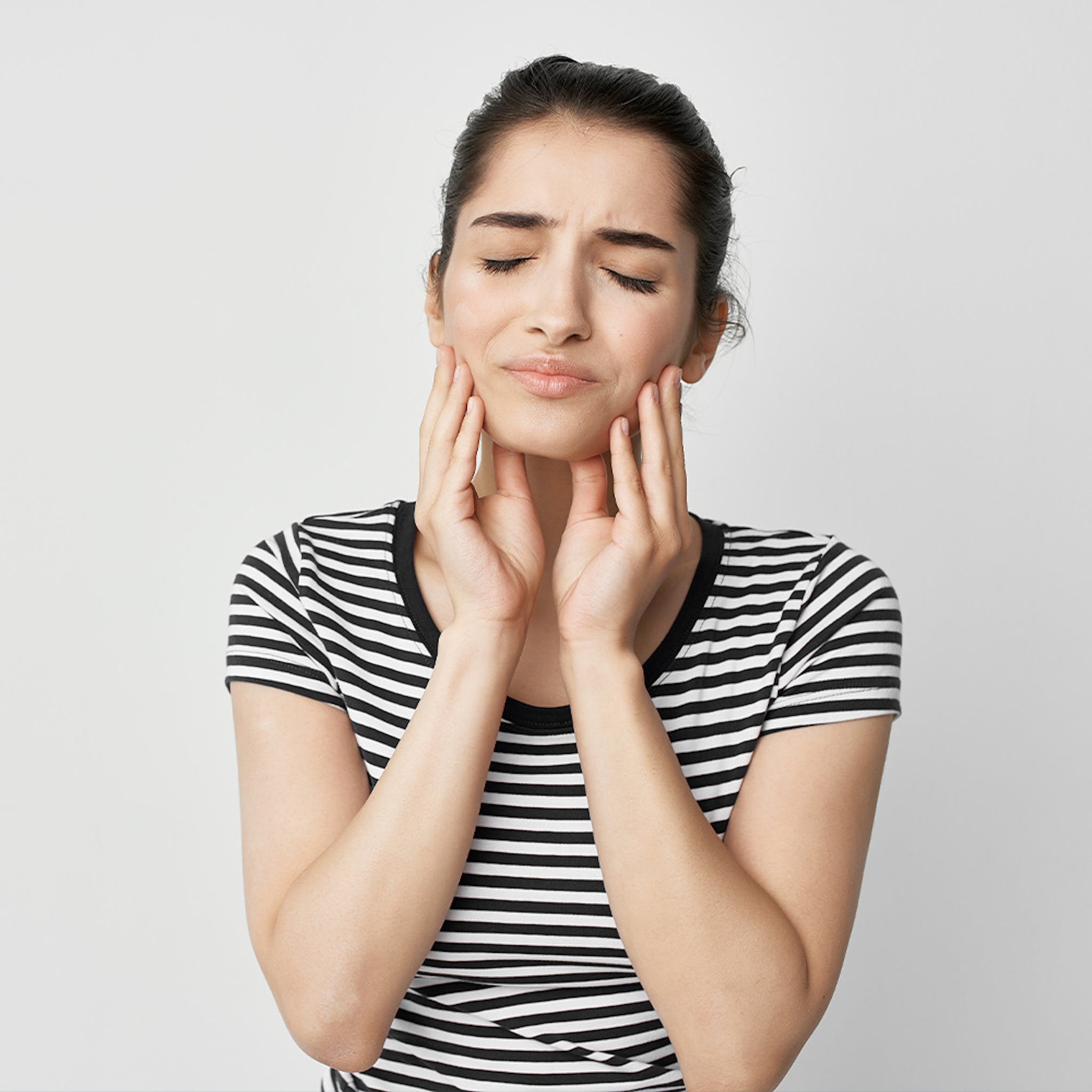 Woman in striped shirt touching face with eyes closed, showing facial discomfort or dental pain.
