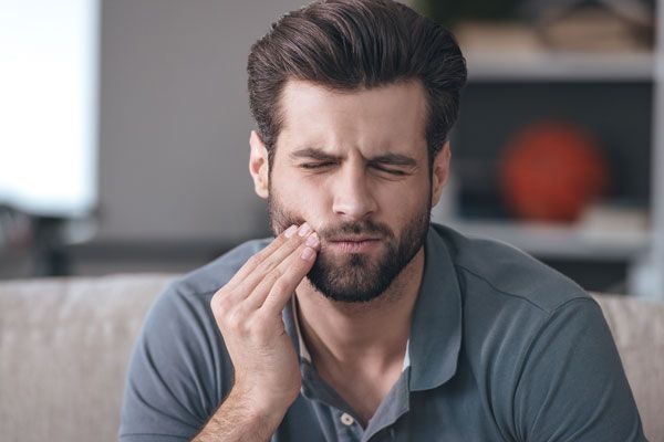 Man with beard sitting on couch, touching face, appearing thoughtful in modern dental office setting.