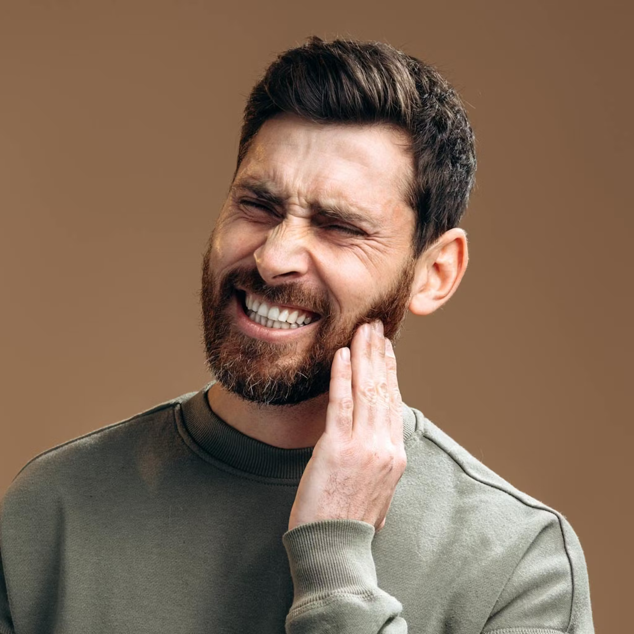 Man smiling while touching his cheek against tan background, dental discomfort concept.