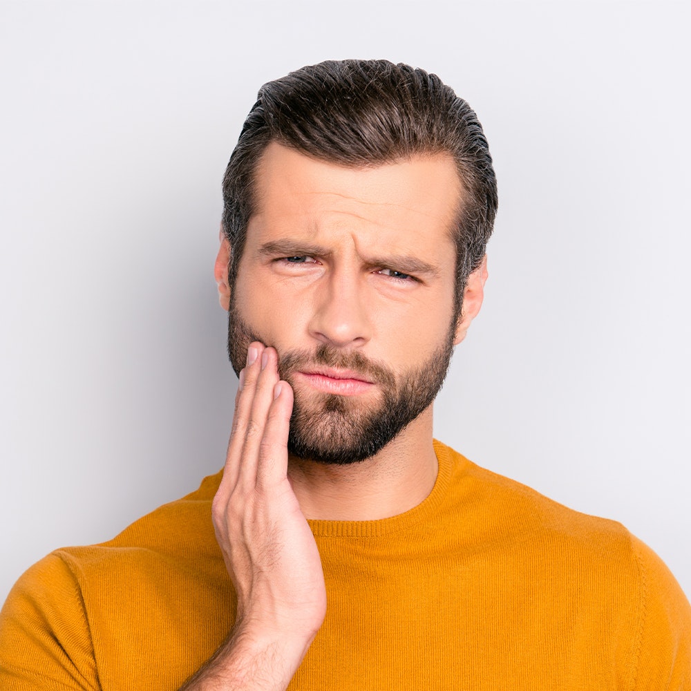 Man in yellow shirt touching his cheek with concerned expression against white background.